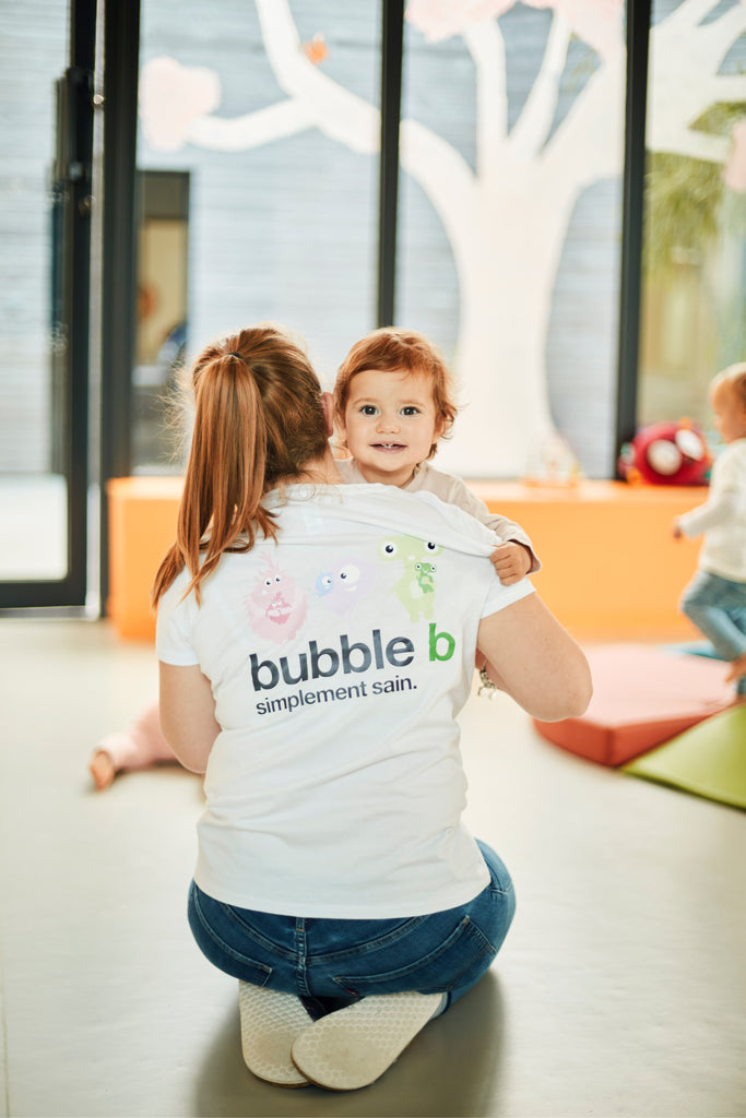 Un bébé souriant dans les bras d'une auxiliaire de puériculture avec un tee-shirt bubble b dans une crèche 